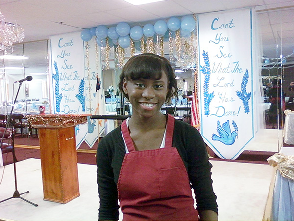 janteyl, a black 15 year old smiles and has her hair pulled back in a pony tail. wearing black shirt and red apron