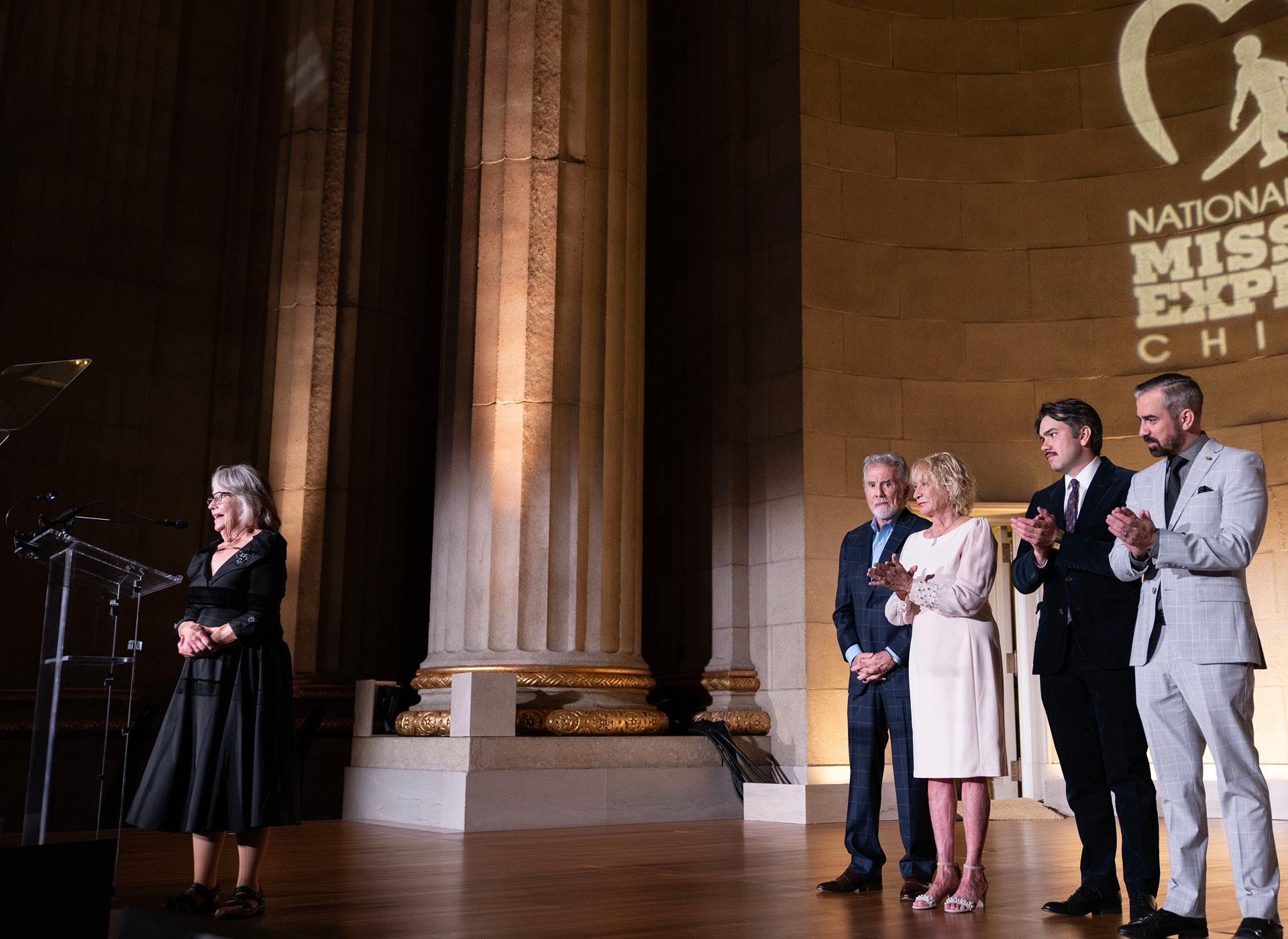 woman in black dress stands at podium as three men in suits and woman in white dress stand behind her