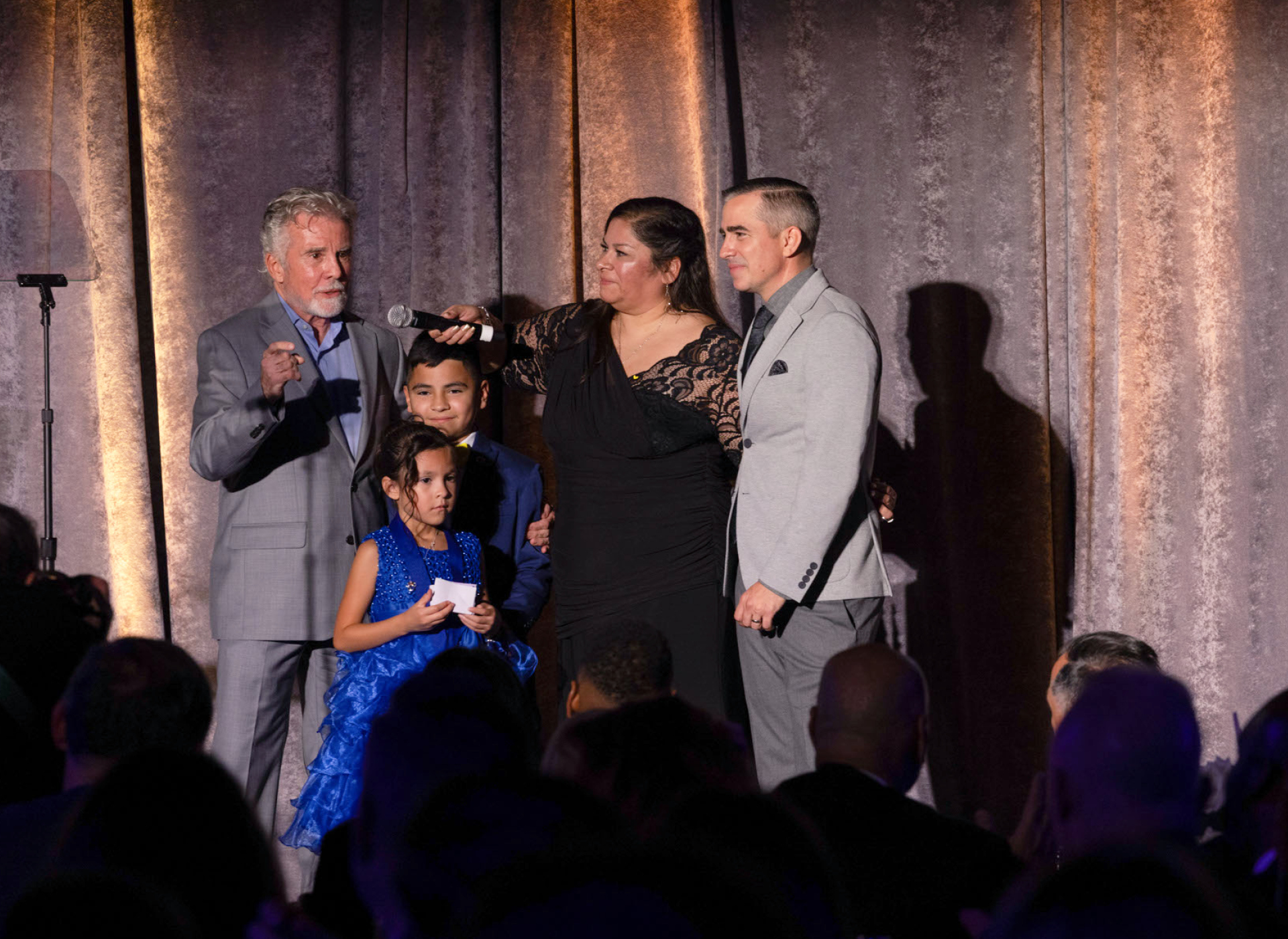 john and callahan walsh both in gray suits stand on stage with woman in black gown, boy in blue suit, and girl in blue dress